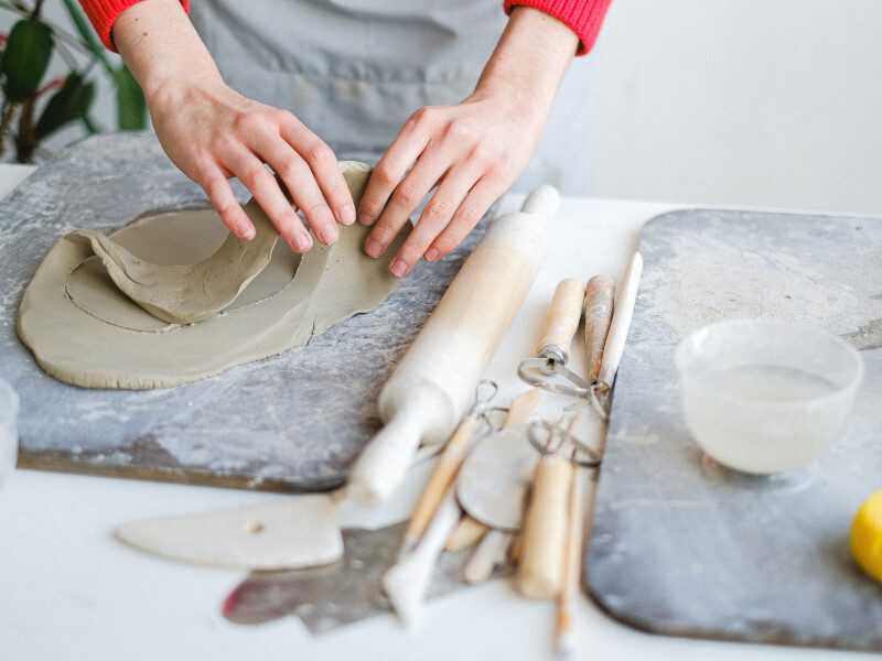 A person stands at a table working on a flat piece of clay with pottery tools next to them